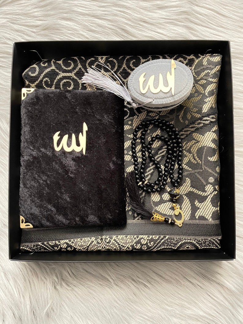 May include: A black gift box containing a prayer mat, a velvet pouch, a prayer bead, and a compass. The prayer mat has a black and gold floral pattern. The pouch and compass feature the word "Allah" in gold.