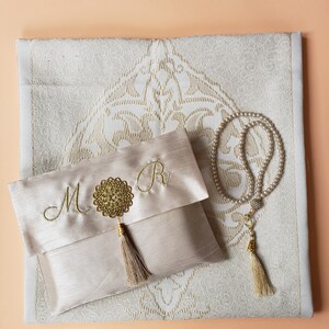 Puede incluir: Una alfombra de oraci&oacute;n beige con un dise&ntilde;o bordado en oro. Una bolsa de sat&eacute;n beige a juego con un borla dorada y las letras "M" y "R" bordadas en ella. Un rosario de oraci&oacute;n de perlas blancas con una borla dorada.