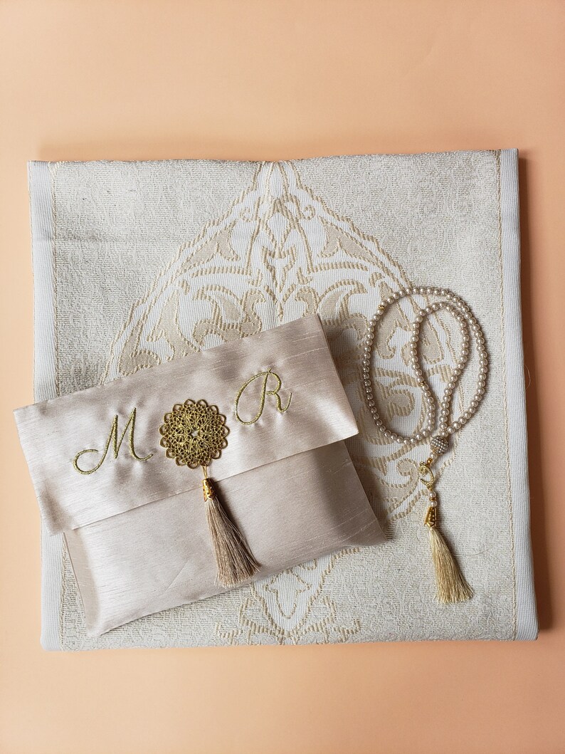 Puede incluir: Una alfombra de oraci&oacute;n beige con un dise&ntilde;o bordado en oro. Una bolsa de sat&eacute;n beige a juego con un borla dorada y las letras "M" y "R" bordadas en ella. Un rosario de oraci&oacute;n de perlas blancas con una borla dorada.