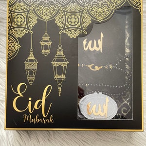 May include: A black gift box with gold detailing, featuring the words "Eid Mubarak". Inside, there's a prayer mat, prayer beads, and a decorative item, all with gold accents. The box has a decorative pattern.