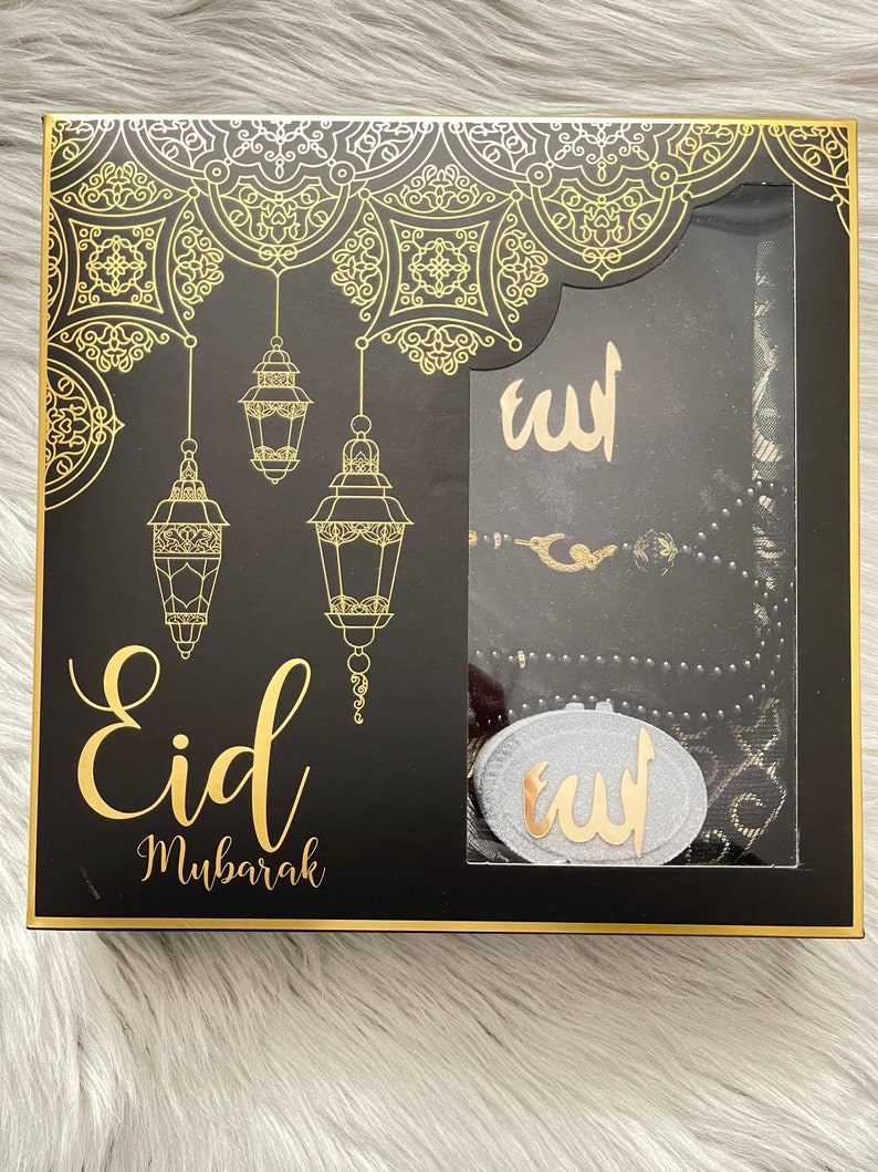 May include: A black gift box with gold detailing, featuring the words "Eid Mubarak". Inside, there's a prayer mat, prayer beads, and a decorative item, all with gold accents. The box has a decorative pattern.