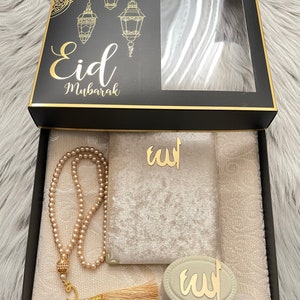 May include: A black gift box with gold detailing, labelled "Eid Mubarak". Inside, a cream-coloured prayer mat, a beaded rosary, a velvet pouch, and a decorative item with Arabic script are visible. The box lid features a decorative design with hanging lanterns.