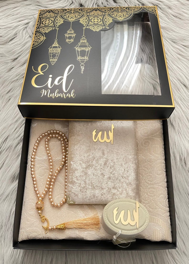 May include: A black gift box with gold detailing, labelled "Eid Mubarak". Inside, a cream-coloured prayer mat, a beaded rosary, a velvet pouch, and a decorative item with Arabic script are visible. The box lid features a decorative design with hanging lanterns.