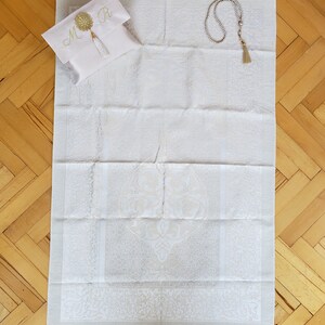 Puede incluir: Alfombra de oraci&oacute;n blanca con un dise&ntilde;o bordado en oro y una borla blanca. Se incluye una bolsa blanca con bordado en oro y las iniciales "M R". Tambi&eacute;n se incluye un collar de cuentas de oraci&oacute;n blanco.