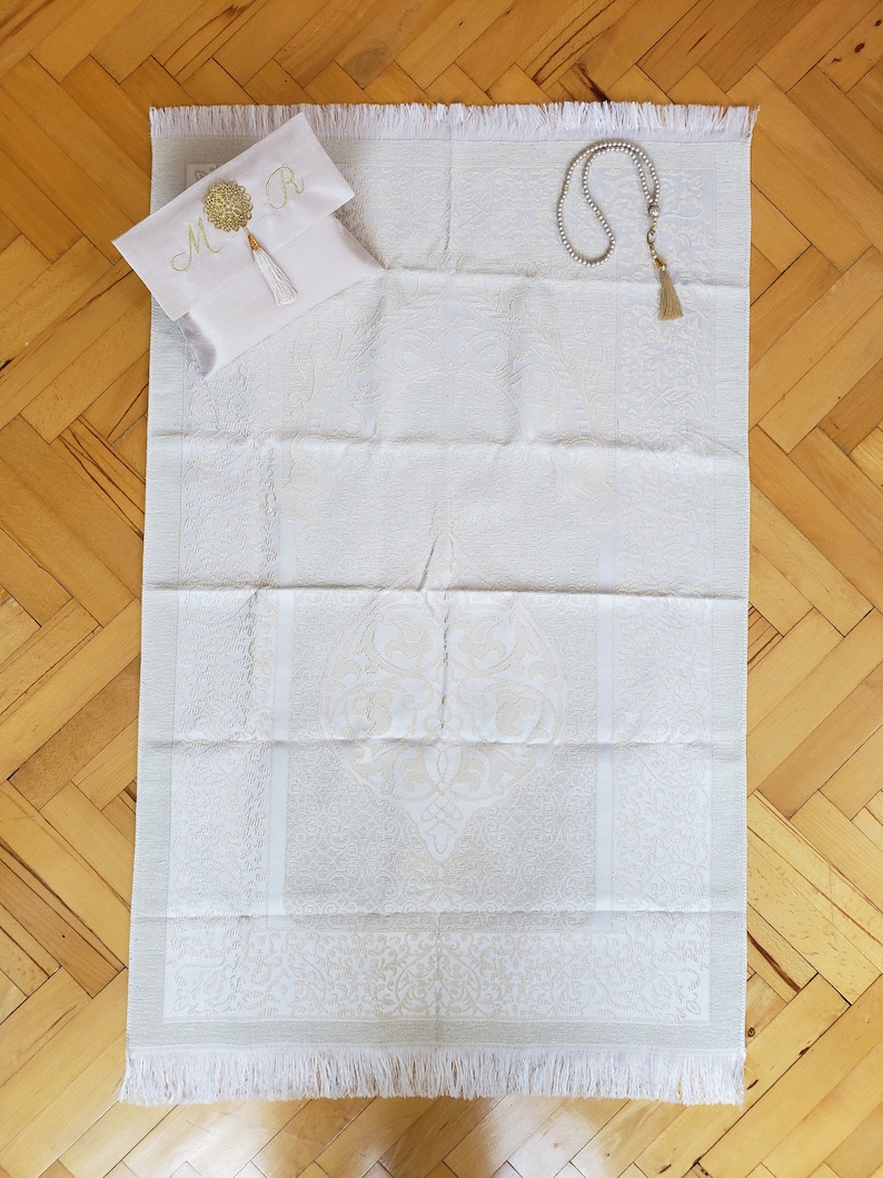 Puede incluir: Alfombra de oraci&oacute;n blanca con un dise&ntilde;o bordado en oro y una borla blanca. Se incluye una bolsa blanca con bordado en oro y las iniciales "M R". Tambi&eacute;n se incluye un collar de cuentas de oraci&oacute;n blanco.