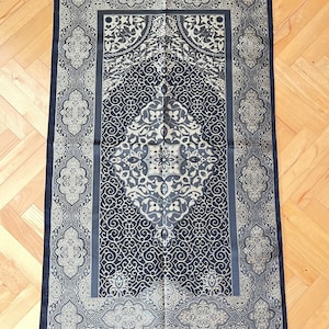May include: A navy blue prayer mat with a detailed, symmetrical pattern in grey and cream. It has a central medallion and decorative borders, with a fringe along the bottom. The design incorporates floral and geometric motifs.
