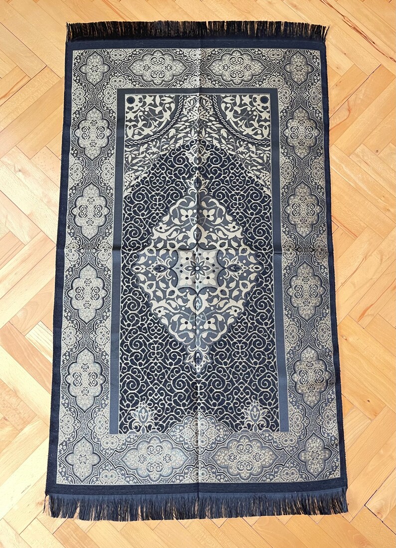 May include: A navy blue prayer mat with a detailed, symmetrical pattern in grey and cream. It has a central medallion and decorative borders, with a fringe along the bottom. The design incorporates floral and geometric motifs.