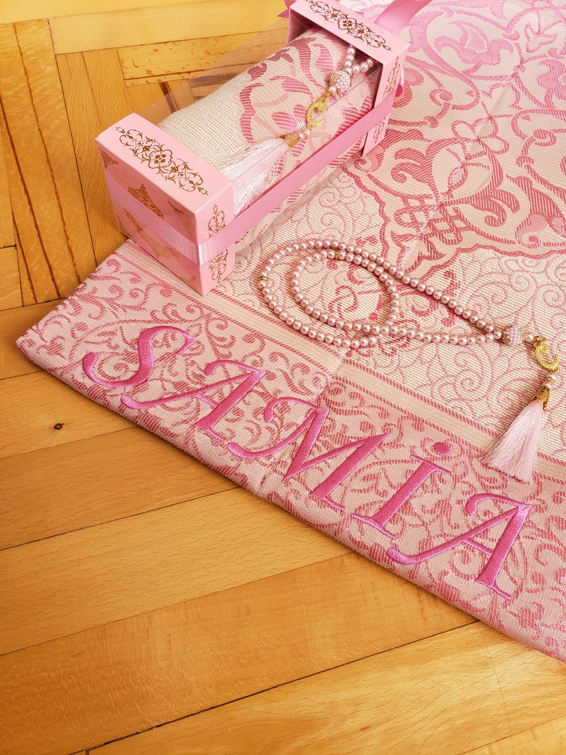 May include: A pink prayer rug with a matching pink box containing a prayer rug and a pink prayer beads. The prayer rug has the name "SAMIA" embroidered on it in pink.