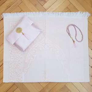 Puede incluir: Una alfombra de oraci&oacute;n rosa con un borde blanco con flecos y una bolsa rosa a juego con una borla dorada. Tambi&eacute;n se incluye un collar de cuentas de oraci&oacute;n rosa.