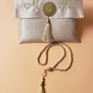Puede incluir: Una bolsa de sat&eacute;n blanco con bordado dorado de las letras "M" y "R" y un borla dorada. Un collar de cuentas de oraci&oacute;n de perlas blancas con una borla dorada y un dije dorado.