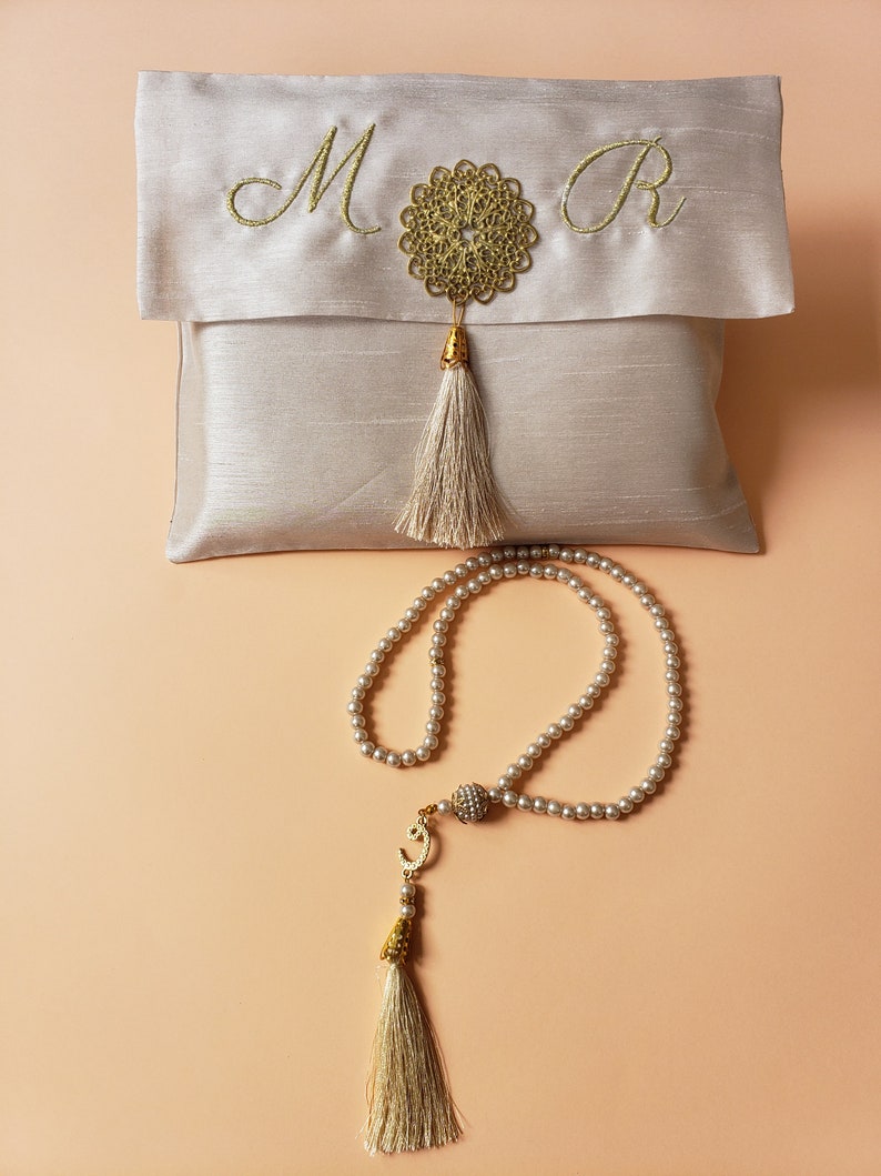 Puede incluir: Una bolsa de sat&eacute;n blanco con bordado dorado de las letras "M" y "R" y un borla dorada. Un collar de cuentas de oraci&oacute;n de perlas blancas con una borla dorada y un dije dorado.