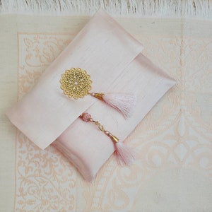 Puede incluir: Una bolsa de sat&eacute;n rosa con un adorno de filigrana dorada y dos borlas rosas. La bolsa est&aacute; sobre una superficie con estampado blanco y rosa.