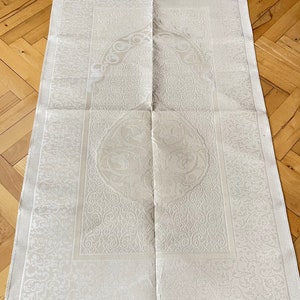 May include: A cream prayer mat with an elaborate floral pattern. The rectangular mat has a decorative border and a fringed edge. The central design features a circular motif. The mat is folded.