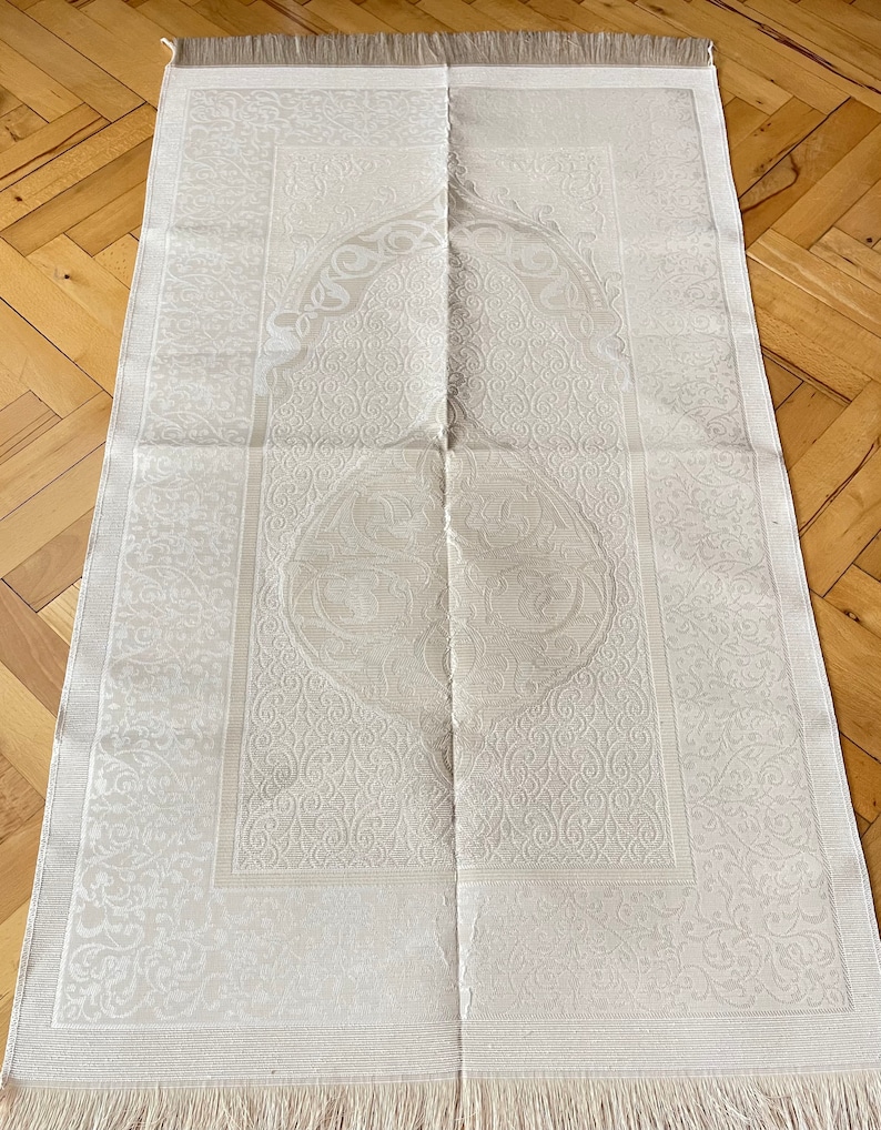 May include: A cream prayer mat with an elaborate floral pattern. The rectangular mat has a decorative border and a fringed edge. The central design features a circular motif. The mat is folded.