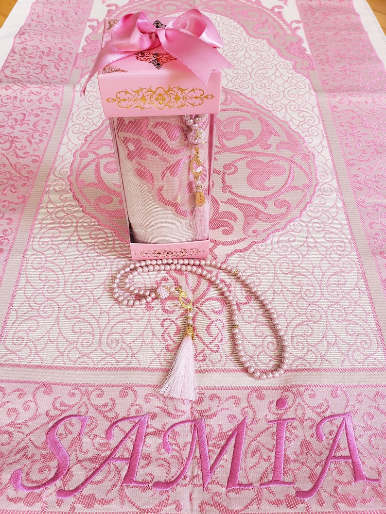 May include: A pink prayer rug with a matching pink prayer beads and a pink box with a gold trim. The rug has the name "SAMIA" embroidered on it.