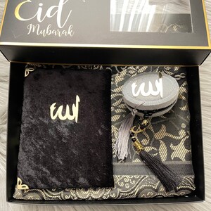 May include: A black gift box with gold edging, containing a black prayer mat, a black velvet book, and a grey and black beaded item. The box lid has the words "Eid Mubarak" in gold script.