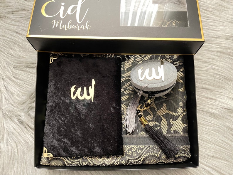 May include: A black gift box with gold edging, containing a black prayer mat, a black velvet book, and a grey and black beaded item. The box lid has the words "Eid Mubarak" in gold script.
