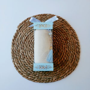 May include: A white scarf with a blue tassel and gold beads is packaged in a light blue box with a gold floral design. The box is tied with a white ribbon and sits on a woven brown mat.