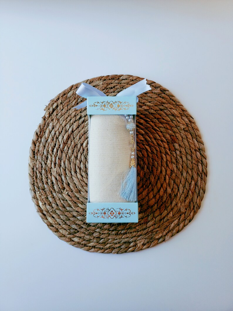 May include: A white scarf with a blue tassel and gold beads is packaged in a light blue box with a gold floral design. The box is tied with a white ribbon and sits on a woven brown mat.