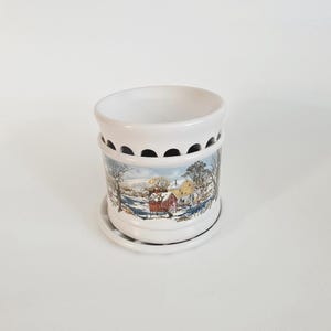 May include: A white ceramic wax warmer featuring a cylindrical body with a scalloped top edge. The exterior is decorated with a colorful winter landscape scene depicting snow-covered trees, a red barn, and a yellow house near a frozen stream.