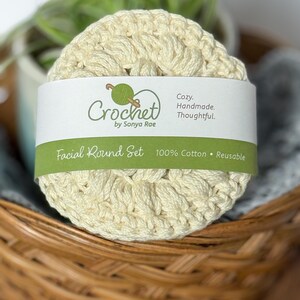 May include: A set of cream-colored, crocheted facial rounds, wrapped with a white band featuring the text "Crochet by Sonya Rae" and "Facial Round Set 100% Cotton • Reusable." The rounds are in a woven basket.