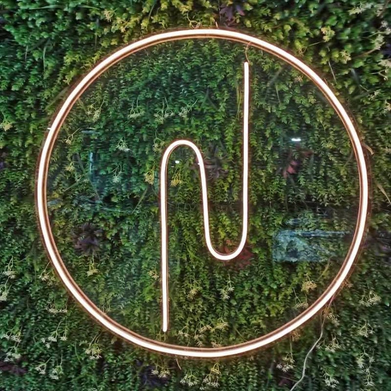May include: A white neon sign in the shape of the letter "N" is displayed in a circle against a green wall of artificial foliage.