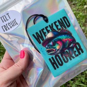 May include: A turquoise blue felt car air freshener with a black and white illustration of a fish wearing sunglasses and a baseball cap. The text "Weekend Hooker" is printed on the air freshener.