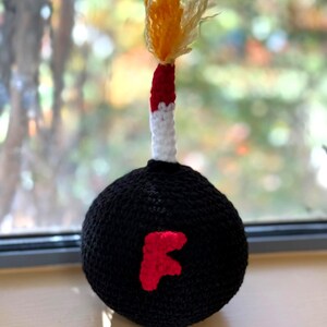 May include: A handmade crochet bomb with a black spherical body and a red "F" applique. The fuse is white, red, and yellow, resembling a lit flame. The craft item is displayed on a light-colored surface.