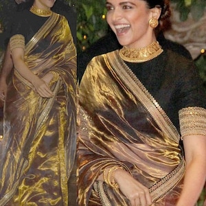 May include: A woman in a black top and a gold sari with detailed embellishments. The sari has a shimmering, metallic finish. She is also wearing gold jewellery, including a necklace and earrings.