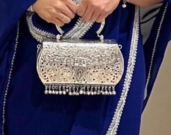 May include: A silver, ornate handbag with intricate filigree detailing and a small handle is held against a backdrop of a deep blue garment. The bag features a decorative clasp and dangling beaded fringe along the bottom edge.