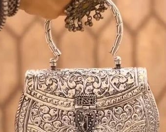 May include: Silver-colored, intricately carved handbag with a curved handle. The bag features detailed floral and geometric patterns, a heart-shaped clasp, and small dangling embellishments. The bag is held by a hand wearing matching silver jewelry.