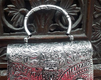 May include: A silver metal clutch purse with an ornate floral design and a red stone accent. The purse has a top handle.