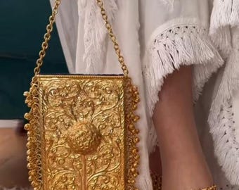 May include: A gold-colored, rectangular handbag with intricate floral and scrollwork designs. The bag features a chain strap and is adorned with small, dangling beads along the edges. A matching gold anklet is also visible.