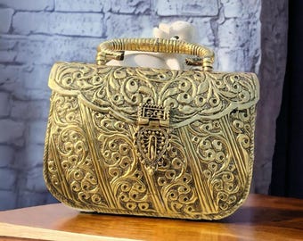 May include: Ornate gold-colored handbag with intricate floral and scrollwork patterns. The bag features a decorative clasp and a textured handle. The design is symmetrical, with vertical and curved lines. The bag is sitting on a wooden surface.