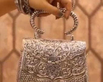 May include: Ornate silver handbag with intricate floral and scrollwork patterns. The bag features a clasp closure and decorative handles. Matching silver bracelets adorn the wrist holding the bag. The overall design is detailed and elegant.