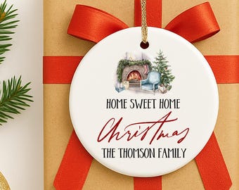 May include: A round white Christmas ornament with a gold string, featuring a cozy fireplace scene and the words "Home Sweet Home Christmas" and "The Thomson Family" in red script. The ornament is on a wrapped gift with a red bow.