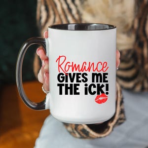 Puede incluir: Taza de cerámica blanca con interior y asa negros. La taza presenta el texto "Romance GIVES ME THE iCK!" en rojo y negro, junto con una huella de labios rojos. La taza está sujeta por una persona.