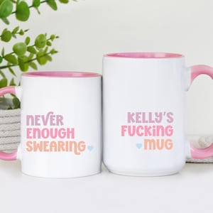 May include: Two white ceramic mugs with pink handles and interiors. One mug reads "NEVER ENOUGH SWEARING" and the other says "KELLY'S FUCKING MUG" in pink and orange text. Both mugs have a small blue heart.