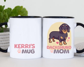 Op de afbeelding: Witte keramische mok met een zwart handvat en rand. De mok heeft de tekst "Kerri's Mug" in roze en paars, en een cartoon teckel met de tekst "Dachshund Mom". De mok heeft een pootafdrukontwerp.