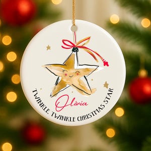 Peut inclure: Une décoration de Noël ronde et crème avec un motif d'étoile dorée. L'étoile a un visage souriant et est ornée d'un nœud rouge. L'ornement porte le nom "Olivia" et le texte "Twinkle Twinkle Christmas Star".