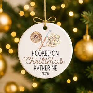 May include: A white ceramic Christmas ornament with a gold string. The ornament features a watercolor illustration of yarn, a crochet hook, and a crocheted flower. Text reads "Hooked on Christmas KATHERINE 2025."