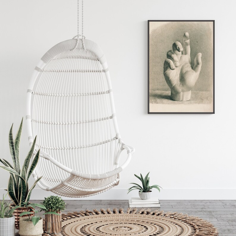 May include: A white wicker hanging chair with a woven design hangs from the ceiling in a room with a white wall and a framed drawing of a hand. The room has a light wood floor and a brown woven rug.