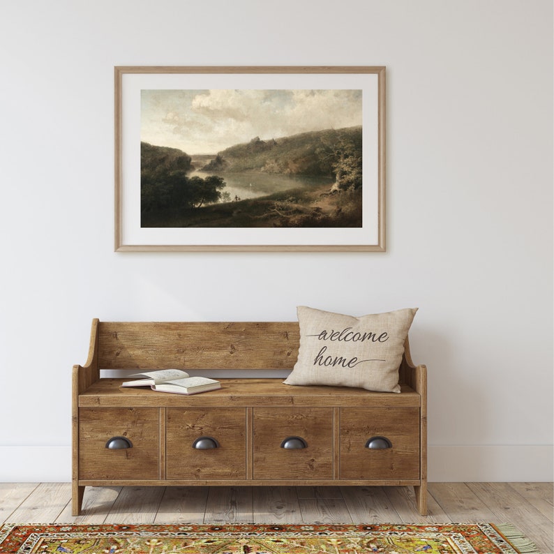 May include: A framed landscape painting hangs above a wooden bench with four drawers. A decorative pillow with the words "welcome home" rests on the bench. An open book sits on the bench.