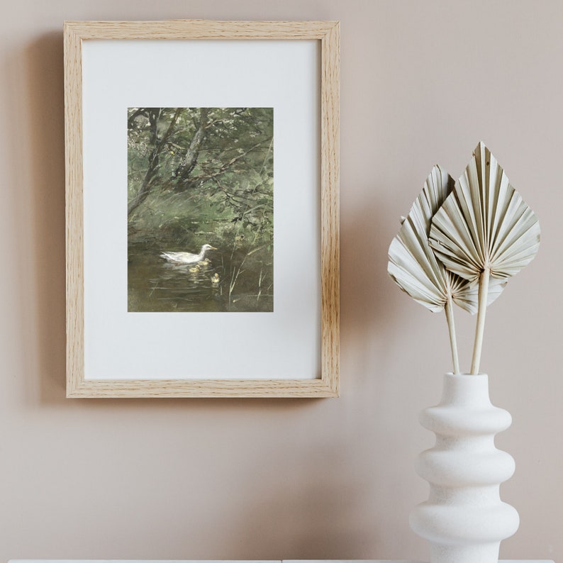Op de afbeelding: Een ingelijste print van een aquarel schilderij met een witte eend die zwemt in een vijver met twee eendjes. De vijver is omgeven door groene bomen en bladeren.