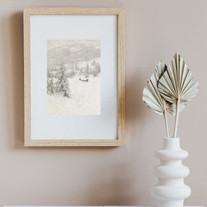 Peut inclure: Une impression encadr&eacute;e d'un paysage d'hiver enneig&eacute; avec une petite silhouette marchant au loin. L'impression est dans un cadre en bois clair et est accroch&eacute;e &agrave; un mur rose p&acirc;le.