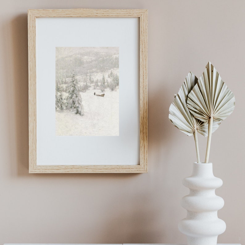 Peut inclure: Une impression encadr&eacute;e d'un paysage d'hiver enneig&eacute; avec une petite silhouette marchant au loin. L'impression est dans un cadre en bois clair et est accroch&eacute;e &agrave; un mur rose p&acirc;le.
