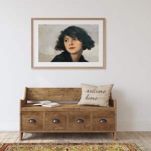 Op de afbeelding: Een ingelijst schilderij van een vrouw met donker haar en een lichtblauwe achtergrond. Het schilderij hangt aan een witte muur boven een houten bank met een kussen waarop "welkom thuis" staat.