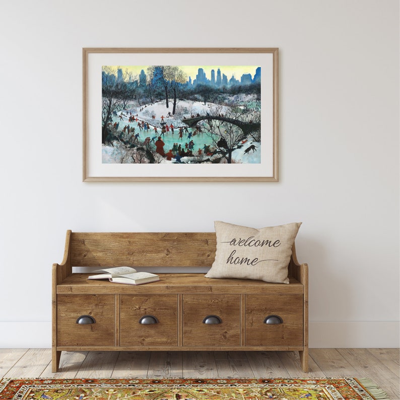 May include: A framed watercolour painting of a winter scene in Central Park, New York City. The painting depicts people ice skating on a frozen pond with a bridge in the background. The cityscape is visible in the distance. The painting is hanging on a white wall above a wooden bench with drawers.