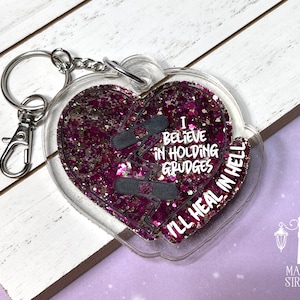 May include: A pink and silver glitter filled heart-shaped keyring with a broken heart design and the text "I believe in holding grudges I'll heal in hell".