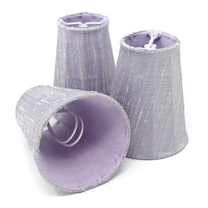 May include: Three small, tapered lampshades in a muted lavender colour are arranged against a white background. The shades have a textured appearance and a lighter lavender interior. Each shade has a metal wire frame.